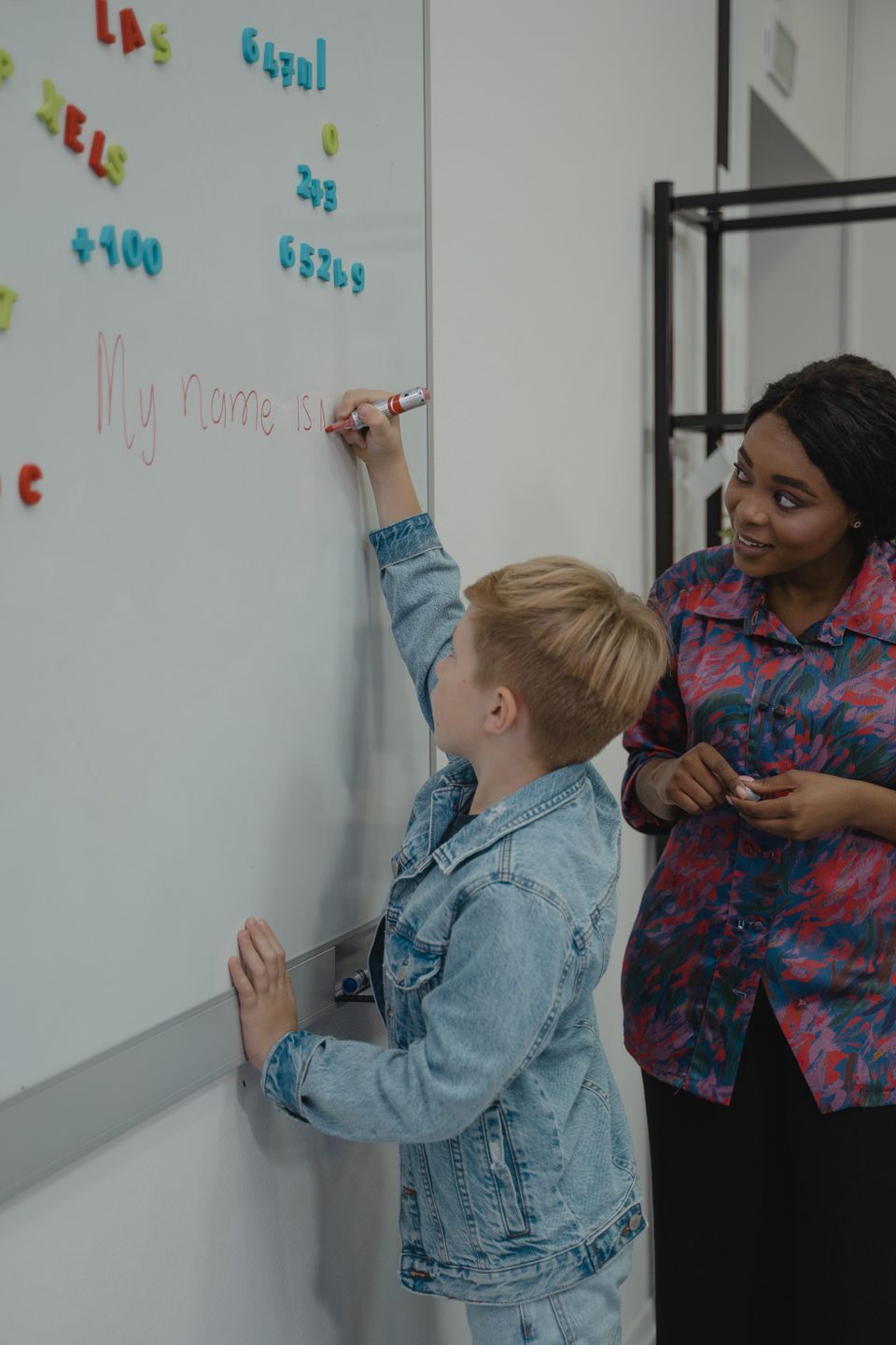 A young student writing "My name is" on a whiteboard with a red marker while a tutor watches